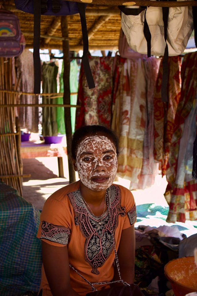 Woman with a traditional face mask in a Malagasy market stall displaying vibrant fabrics.